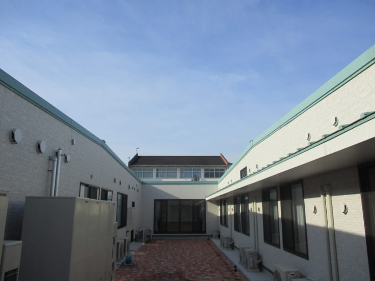 介護施設の建物外観。中庭を囲む複数棟の白い建屋と緑色屋根が見えます。